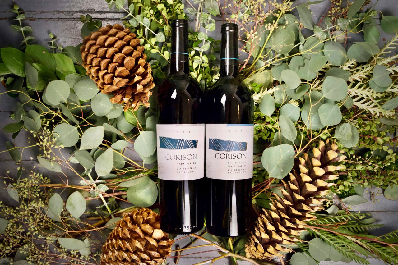 A bottle each of the 2011 and 2021 Corison Napa Valley Cabernet Sauvignons surrounded by pine cones and holiday greenery.