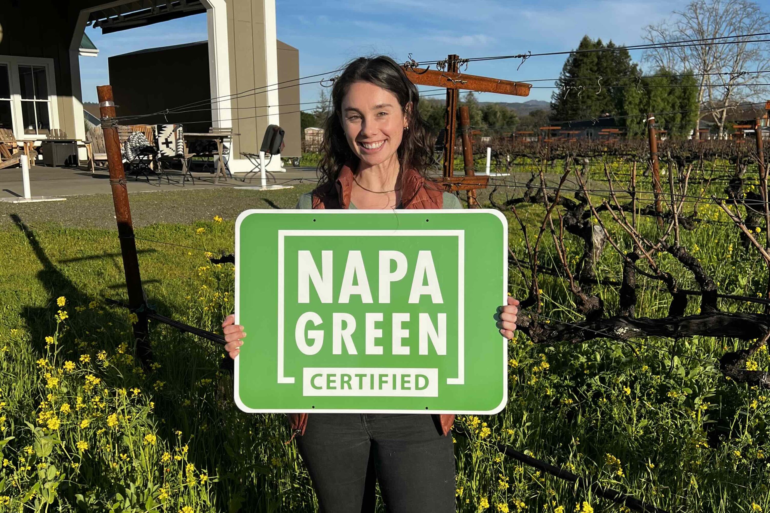 Grace Corison Martin holding the "Napa Green Certified" sign in Kronos vineyard, with Corison winery in the background.